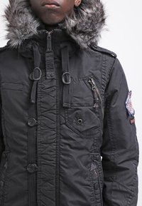 Parka nera con cappuccio in pelliccia sintetica, chiusura a zip e bottoni, numerose tasche con zip e bottoni, e texture trapuntata.
