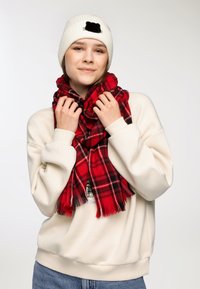 Czerwona kraciasta szalik z frędzlami noszony na kremowej bluzie. Wełniana biała czapka mały czarny detal.