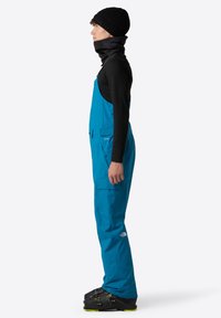 Blauwe waterdichte overalls met meerdere zakken, verstelbare banden en versterkte naden. Draagbaar met een zwarte beanie en nekwarmer.
