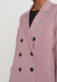 Jasnofioletowa marynarka dwurzędowa z gładkiego materiału. Wyposażona w czarne guziki, dwie boczne kieszenie oraz kołnierz z wycięciem.