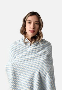 Manta suave y texturizada en un patrón de rayas azul y crema, con acentos de bobble en relieve. Usada cómodamente alrededor de los hombros.