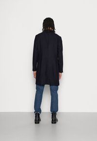 Hombre con trenzas se encuentra de frente a la pared, vistiendo un largo abrigo negro, jeans azules y botas negras sobre un suelo gris.
