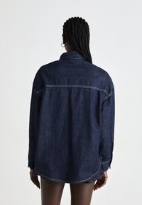 Denim shirt in dark blue with long sleeves, button cuffs, and a curved hem. Features visible stitching and a classic collar.