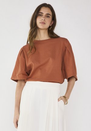 Donna che indossa una blusa color ruggine a maniche corte con spalle arricciate e una gonna bianca plissettata, accessoriata con orecchini a cerchio dorati e braccialetti.