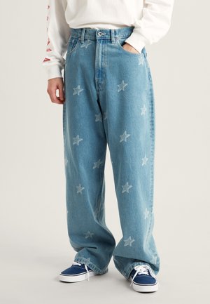 Jeans in denim azzurro chiaro con vestibilità rilassata, caratterizzati da un motivo a stelle in una tonalità più chiara. Abbinati a una camicia bianca a maniche lunghe e sneakers blu navy.