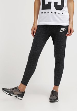 Osoba nosząca czarne spodnie dresowe Nike i czarne sportowe buty, stojąca z jedną nogą lekko ugiętą na jednolitym tle.