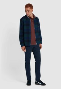 Giacca a quadri blu navy in tessuto morbido con dettagli bianchi, indossata sopra un polo bordeaux, abbinata a pantaloni scuri e sneakers nere.