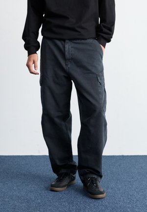 Mand iført løse sorte cargobukser med sidelommer, sorte sneakers med brune såler og en sort langærmet top, stående på en blå tæppe.