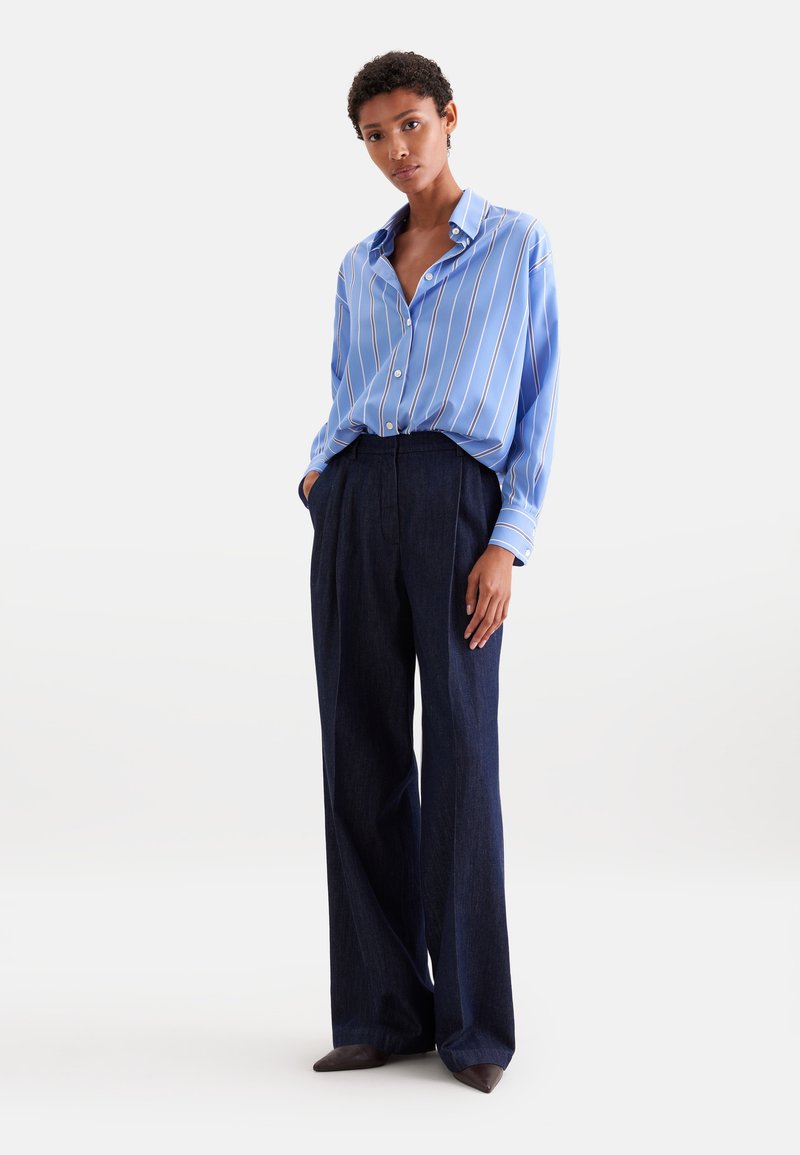 Femme debout portant une chemise bleue rayée ample rentrée dans un pantalon large sombre taille haute et des chaussures pointues foncées sur un fond uni.