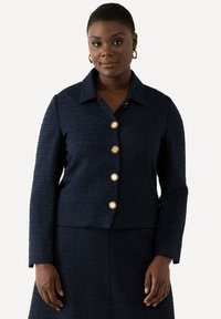 Giacca blu navy con texture e colletto, con sei bottoni dorati sul davanti. Maniche lunghe e vestibilità aderente.