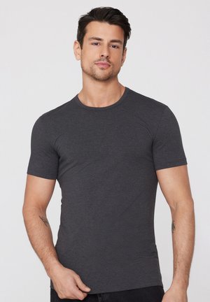 Mann trägt ein eng anliegendes, dunkelgraues T-Shirt mit kurzen Ärmeln und hat Tätowierungen auf beiden Unterarmen. Er steht vor einem schlichten hellen Hintergrund.