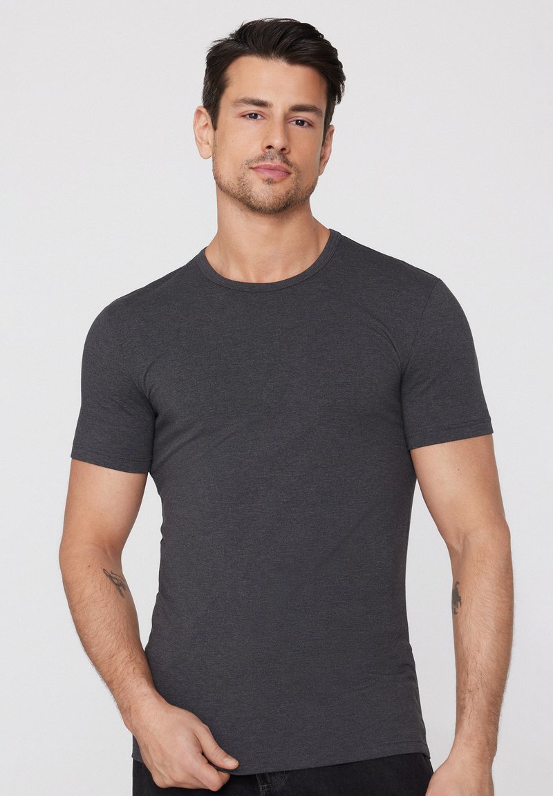 Homme portant un t-shirt gris foncé ajusté à manches courtes avec des tatouages sur les deux avant-bras, debout devant un fond clair uni.