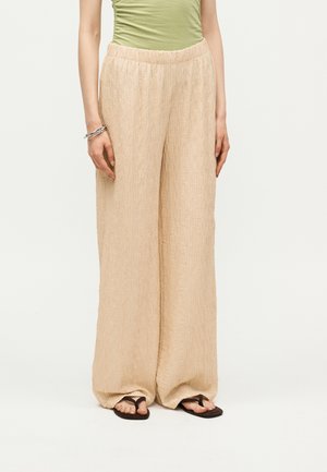 Γυναίκα που φοράει μπεζ υφασμάτινη παντελόνα με φαρδιά πόδια, πράσινο αμάνικο τοπ, ασημένιο βραχιόλι και μαύρα σαγιονάρα, στέκεται μπροστά σε ένα μονόχρωμο φόντο.