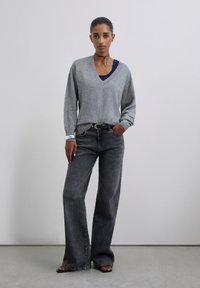 Žena stojí proti šedé stěně, na sobě má šedý svetr s výstřihem do V přes námořnický top, široké vyprané černé džíny, černý pásek, černé sandály, choker a náramek.