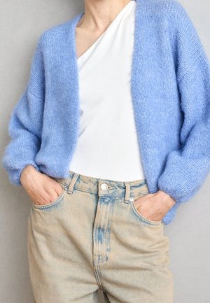 Person trägt eine hellblaue, flauschige Strickjacke über einem weißen, einseitig schulterfreien Top, mit hellbeigen, hoch taillierten Jeans und hat die Hände in den Taschen.