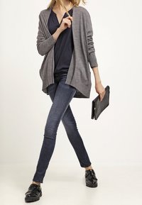 Un cárdigan de punto gris sobre una blusa azul marino, combinado con jeans oscuros y mocasines negros. El modelo sostiene un bolso de mano negro, presentando un atuendo casual.