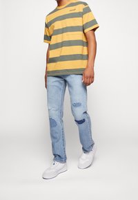 Žluté a šedé proužkované tričko s krátkým rukávem a přední kapsou, spárované s světle modrými roztrhanými džíny a bílými teniskami.