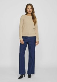 Bežani teksturirani pulover s rebrastim manžetama, uparen s tamnoplavim širokim trapericama i crnim balerinkama s oštrim vrhom, na jednostavnoj bijeloj pozadini.