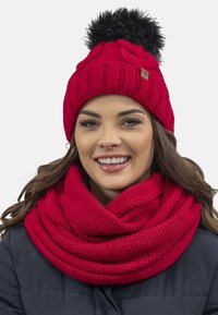Berretto a maglia rosso con un pom-pom nero, caratterizzato da un motivo testurizzato. Accompagnato da una sciarpa infinita rossa a maglia abbinata con una finitura liscia.