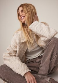 Texturerad krämfärgad jacka med kraag, vävt mönster och framträdande rullade ärmar, kombinerad med ljusbruna jeans med hög midja.