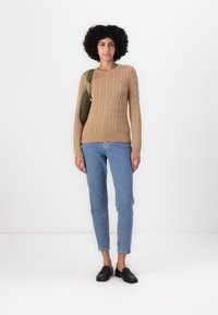 Pull en maille torsadée marron clair avec des manches longues, associé à un jean en denim bleu et des mocassins noirs. La personne porte un sac vert.