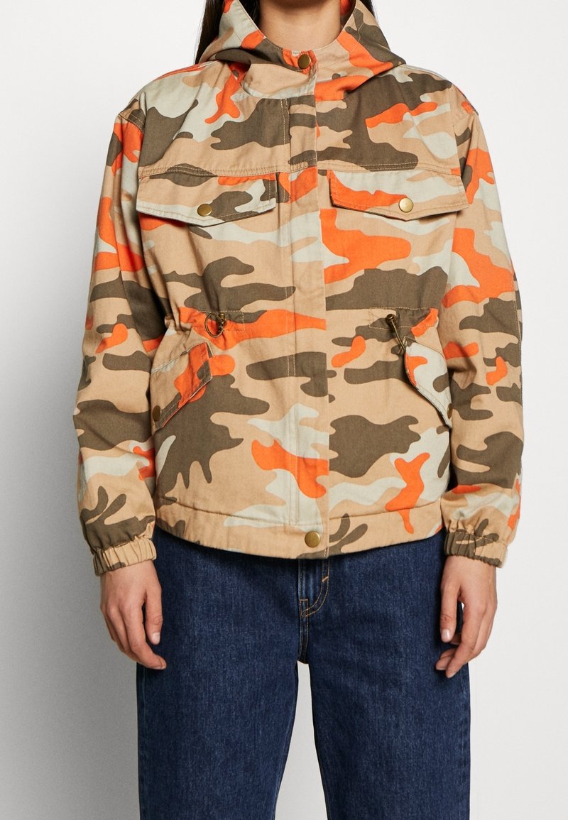 Persona con chaqueta con capucha de camuflaje en tonos beige, marrón, naranja y crema, con bolsillos frontales con solapa y jeans azules visibles desde la cintura hacia abajo.