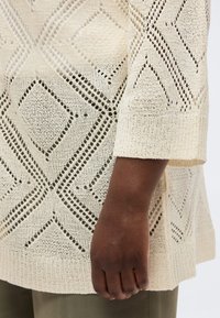 Σκούρο δέρμα χέρι δίπλα σε εκρού πλεκτό πουλόβερ με γεωμετρικά μοτίβα ρόμβου και μανίκια τριών τετάρτων.