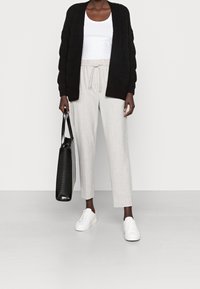 Cardigan nero lavorato a maglia, canottiera bianca, pantaloni grigi chiari con cordino, sneakers bianche, tiene in mano una borsa a tracolla nera con texture.