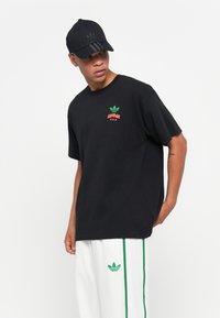 T-shirt en coton noir avec un imprimé de logo Adidas vert et rouge sur la poitrine, associé à un pantalon de survêtement blanc avec des rayures vertes.