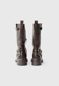 Botas de tobillo de cuero marrón con un acabado texturizado, cremalleras laterales y múltiples correas con herrajes en tono plateado. Tacones cuadrados.