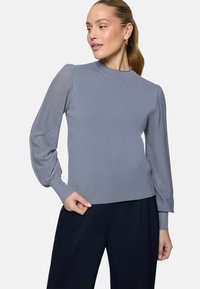 Femme portant un pull en maille bleu clair avec des manches bouffantes texturées et un pantalon noir, regardant sur le côté sur un fond uni.