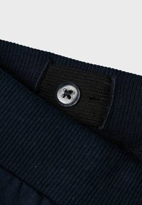 Fascia elastica blu navy con texture a coste, dotata di un bottone di plastica trasparente e un'etichetta nera in tessuto per le informazioni su dimensioni e cura.