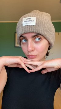 Beanie tricotat bej cu un etichetă rectangulară pe care scrie "MADS NØRGAARD COPENHAGEN." Modelul pozează cu bărbia sprijinită în palme, purtând o cămașă neagră.