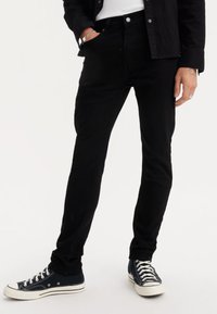 Melnas džinsa bikses ar slaidu piegriezumu, piecām kabatām un metāla pogas aizdari. Kombinētas ar tumši ziliem auduma sporta apaviem.