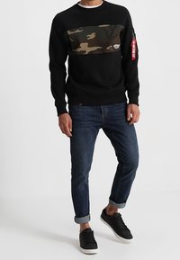 Felpa nera con una striscia camouflage sul petto, dotata di una tasca con zip sul maniche destro e polsini a coste, abbinata a jeans scuri e sneakers nere.