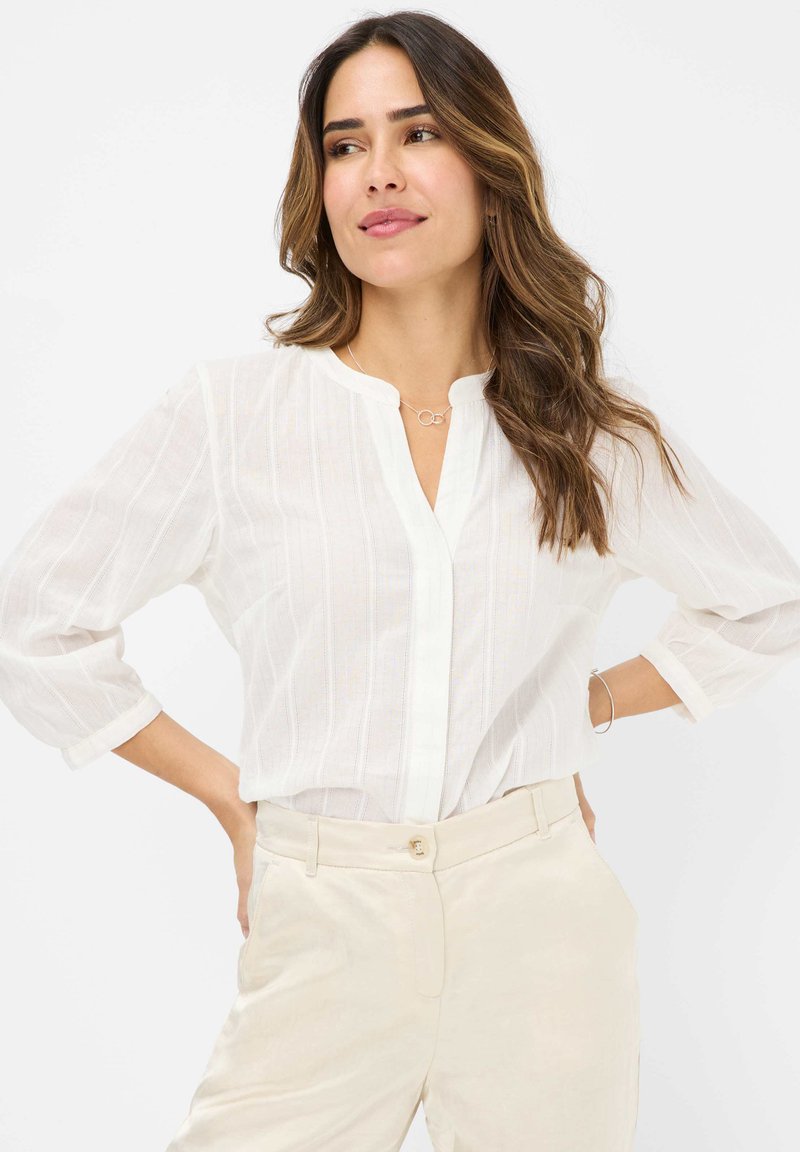 Blusa bianca a maniche lunghe con chiusura a bottoni sul davanti e sottili righe verticali. Il tessuto sembra leggero e leggermente testurizzato.