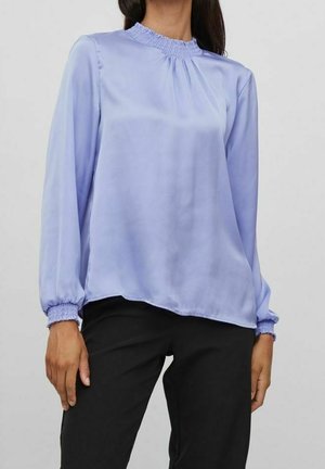 Femme portant un chemisier à manches longues violet clair avec poignets et encolure froncés, associé à un pantalon noir, debout devant un fond uni.