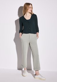 Mörkgrön, texturerad, långärmad cardigan med v-hals och knappar, ihop med ljusgrå, löst sittande byxor och vita sneakers.