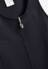 Schwarzes Stofftanktop mit glatter Textur, ausgestattet mit einem Reißverschluss vorne und einem silbernen Zipper in Form eines Löwen. Pflegeetikett im Inneren angenäht.