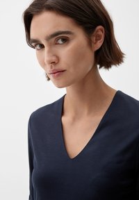 Marineblaue Bluse mit V-Ausschnitt, aus glattem Stoff gefertigt. Hat eine figurbetonte Silhouette und lange Ärmel. Einfacher und minimalistischer Stil.