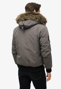 Grijze gewatteerde jas met een met bont gevoerde capuchon, voorzien van zwarte geribde manchetten en een logopatch op de bovenarm. Glad stof, casual pasvorm.