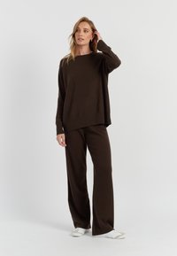 Maglione in maglia marrone con una vestibilità ampia e maniche lunghe, abbinato a pantaloni larghi marroni coordinati. Modella indossa sneaker bianche.