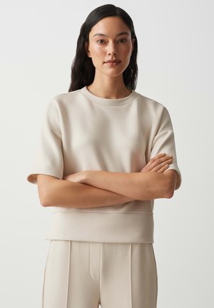 Frau mit langen dunklen Haaren, die ein beiges Kurzarmoberteil und passende Hosen trägt, steht mit verschränkten Armen vor einfachem Hintergrund.