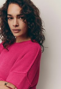 Pulover tricotat roz aprins cu un model texturat, croială lejeră și mâneci scurte. Modelul are părul ondulat închis la culoare și poartă un inel simplu.