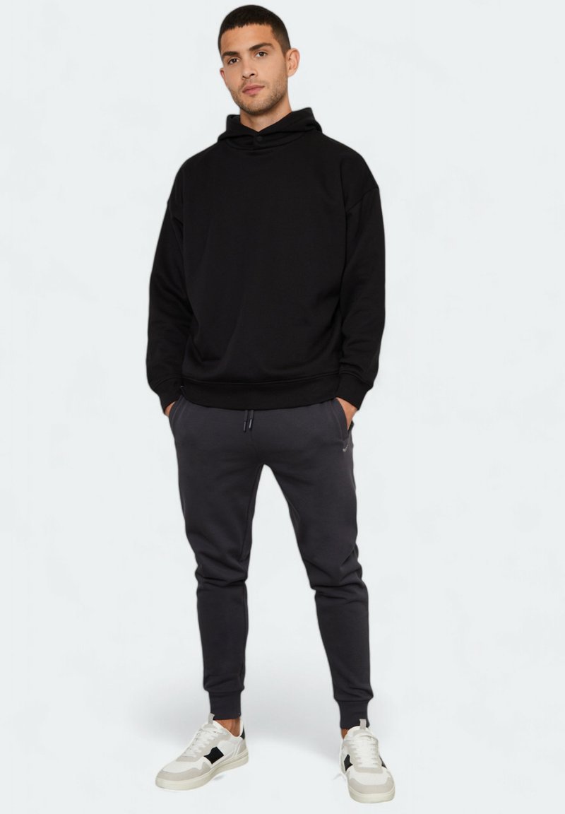 Schwarzer Kapuzenpullover mit lockerer Passform, kombiniert mit dunkelgrauen, schmal zulaufenden Jogginghosen und weißen Sneakers mit schwarzen Akzenten.