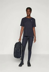 Černé sportovní tričko s krátkými rukávy, doplněné o černé legíny s vzorem. Černý batoh přehozený přes rameno, doplněný o černé tenisky.