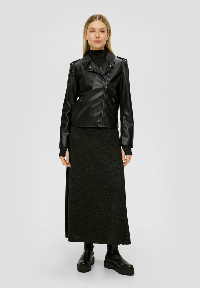 Schwarze Kunstleder-Bikerjacke mit Reißverschluss, Schulterklappen und langen Ärmeln, kombiniert mit einem langen, strukturierten schwarzen Rock und klobigen Stiefeln.