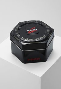 Black hexagonal metal tin with a glossy finish. Features a G-SHOCK logo and embossed text, including "SHOCK RESIST" and "WR 20 BAR."