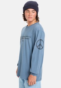 Camiseta de manga larga azul en tejido suave con cuello redondo, que presenta un texto negro y un gráfico de un símbolo de paz en la manga izquierda.