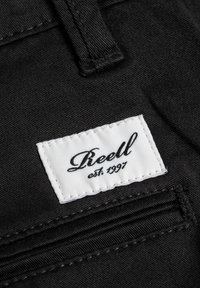 Tela de mezclilla negra con una etiqueta blanca que muestra "Reell est. 1997" en una fuente cursiva, cosida con hilo negro, exhibiendo un diseño limpio.